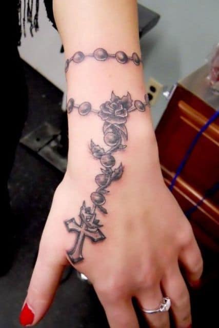 Tatuagem de um terço no braço de uma mulher. O terço começa no antebraço e termina com uma cruz na altura dos dedos 