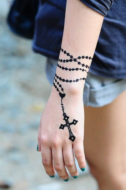 Tatuagem de terço no braço de uma mulher. A corrente começa no antebraço e vai até a altura dos dedos, onde termina com uma cruz. 