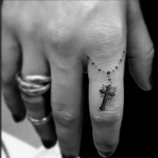 Tatuagem religiosa delicada no dedo de uma mulher com o desenho de uma corrente e uma cruz detalhada na ponta. 