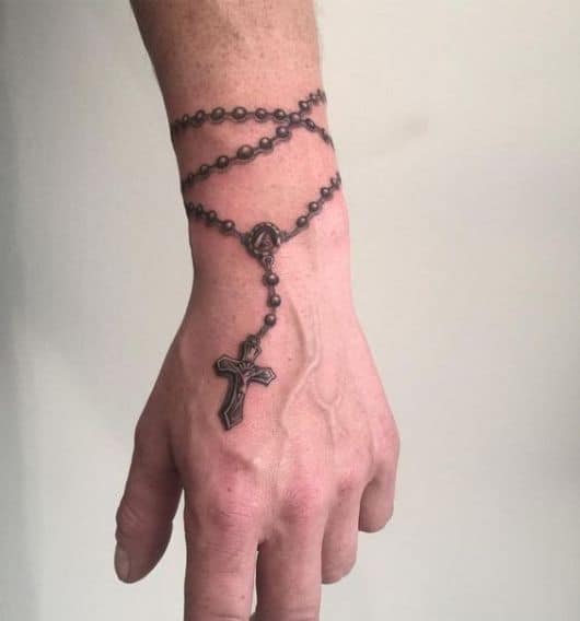 Tatuagem de um terço que começa na altura do pulso de um homem e termina em sua mão com uma cruz. 