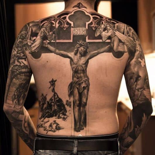 Tatuagem cobrindo as costas de um homem com o desenho de Jesus preso em uma cruz com um anjo de um lado e um demônio do outro. 
