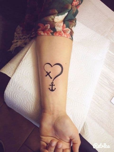 Tatuagem religiosa delicada no antebraço de uma mulher com o desenho de um coração; uma ponta dele forma uma âncora e a outra ponta forma uma cruz. 