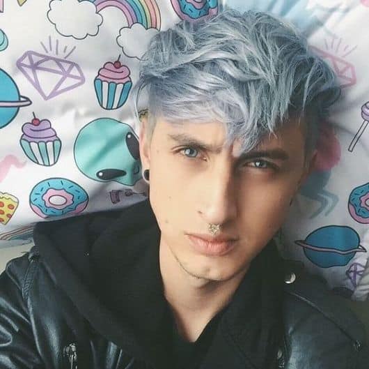 Homem deitado em uma cama tirando uma selfie. Ele tem uma franja pintada com azul platinado. 