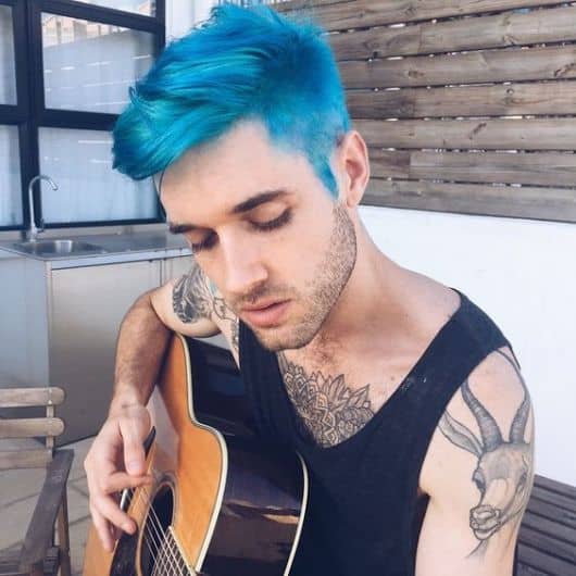 Homem tatuado tocando violão. Seu cabelo é raspado nas laterais com o topo mais longo, toas as partes estão na mesma tonalidade.