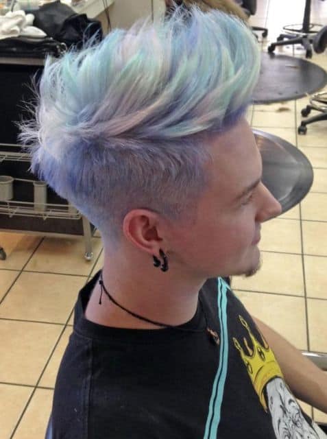 Homem no barbeiro de perfil. Seu cabelo tem um topete grande e é todo pintado com azul platinado, com algumas variações de tons como verde e rosa. 