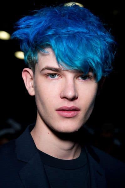 Homem olhando para a câmera vestindo trajes sociais. Seu cabelo é bagunçado com uma franja, quase inteiro pintado de azul com exceção das laterais raspadas. 