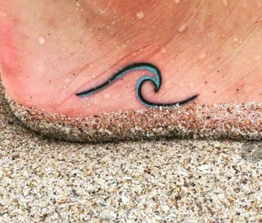 Pé na areia com uma tatuagem no calcanhar de uma onda formada por uma linha colorida de azul na parte interna. 