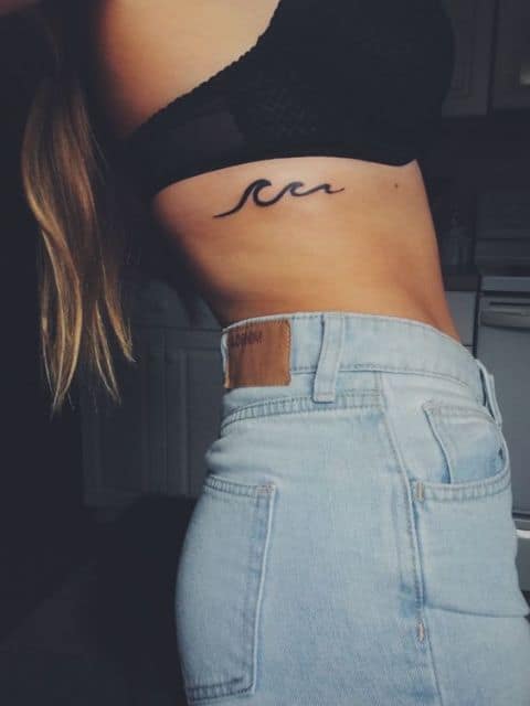 Mulher de biquíni e short jeans. Em sua costela há uma tatuagem com o desenho de três ondas formadas a partir da mesma linha.  