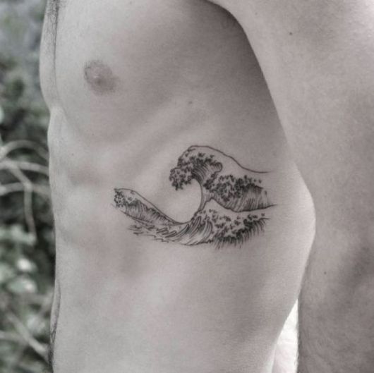 Homem sem camisa com uma tatuagem de onda na costela. A tatuagem é em preto e branco com o desenho de várias ondas se formando no mar. 