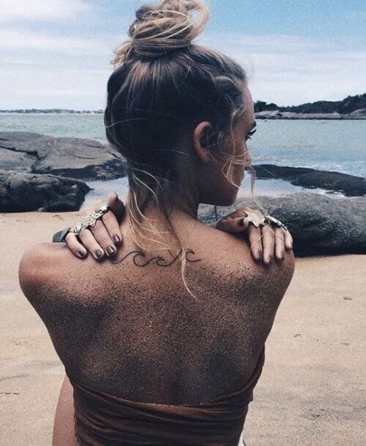 Mulher sentada na areia da praia. No centro de suas costas há uma tatuagem pequena formada por apenas uma linha com o desenho de três pequenas ondas. 