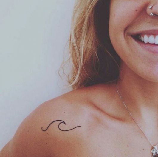Tatuagem pequena de uma onda feita na parte frontal do ombro de uma mulher. 