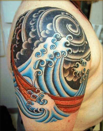 Tatuagem grande no ombro de um homem com o desenho de ondas agitadas engolindo um barco e um céu nublado ao fundo. A tattoo segue uma estética oriental. 