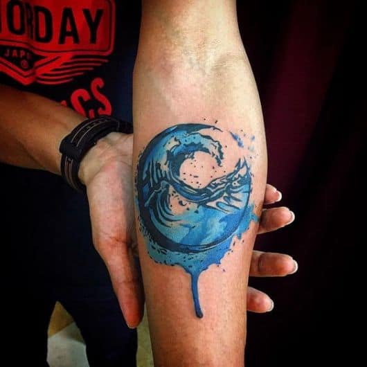 Tatuagem colorida de uma onda dentro de um círculo com uma pequena gota de aquarela escorrendo de dentro dele. 