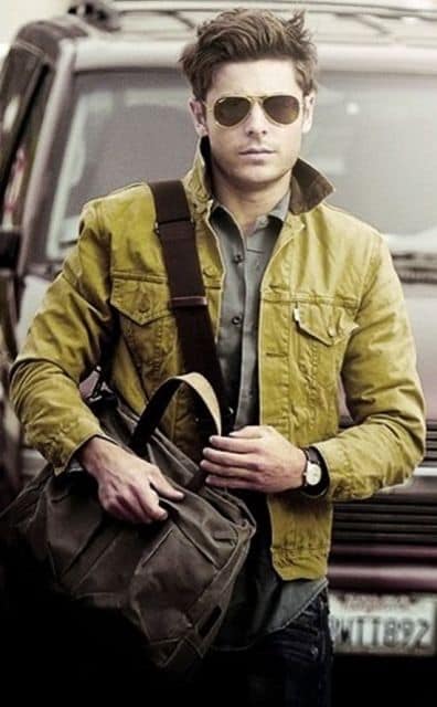 Zac Efron andando na rua. Ele está vestindo uma jaqueta aberta, camisa, calça jeans, relógio, uma bolsa lateral e óculos aviador escuros da Ray Ban. 