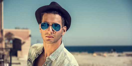 Homem de chapéu em uma praia com o fundo desfocado usando um óculos aviador com lente espelhada levemente azul. 