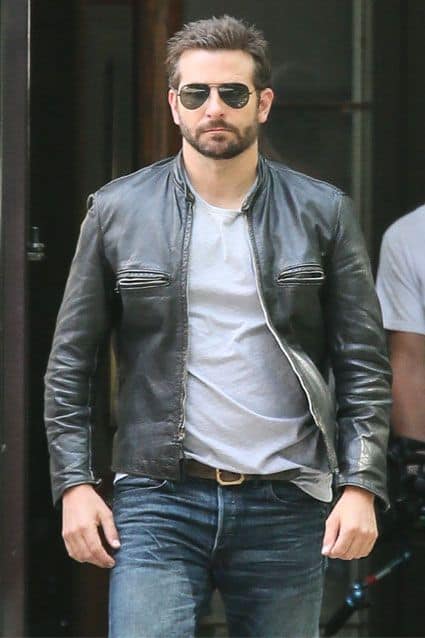 Bradley Cooper andando na rua. Ele está vestindo uma jaqueta de couro, camiseta lisa, calça jeans e um óculos aviador escuro. 