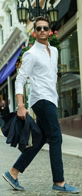 Homem andando na rua vestindo trajes sociais. Ele segura seu terno na mão e os botões superiores de sua camisa estão abertos. Ele também usa um óculos aviador escuro. 