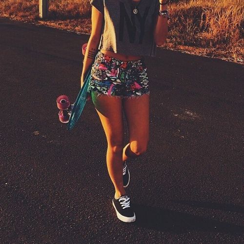 Menina com shorts florido, skate e tênis preto.
